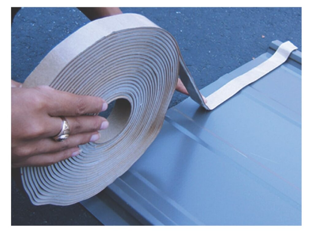 Hands unrolling a roll of butyl tape onto a gray metal roof panel